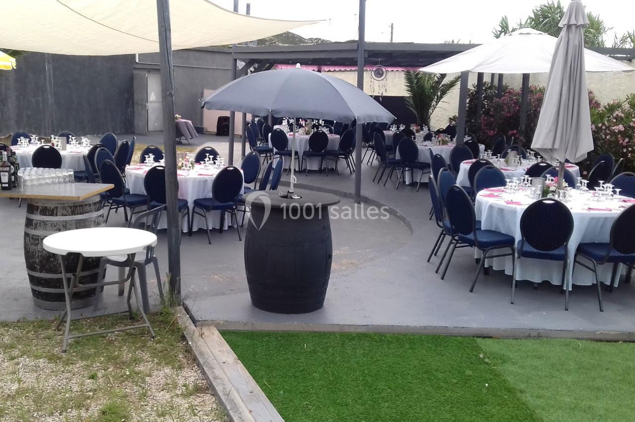 Terrasse aménagée avec des tables rondes dressées, des chaises bleues et des parasols, entourée de végétation.