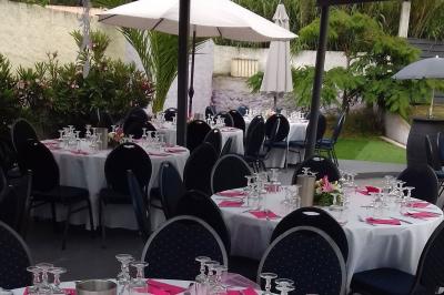 Tables rondes dressées avec nappes blanches, serviettes roses et verres, disposées en extérieur sous des parasols.
