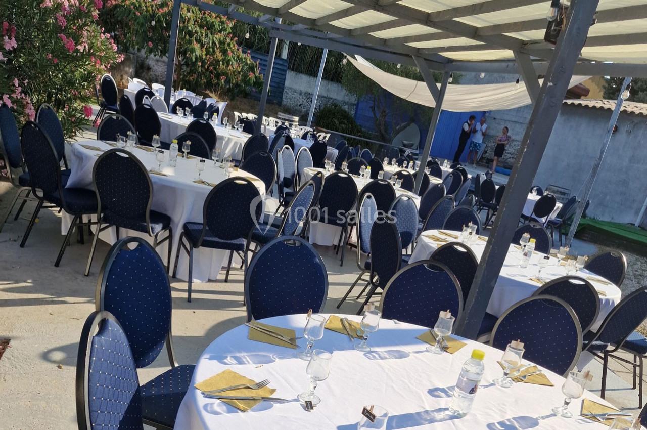 Tables rondes dressées avec nappes blanches et chaises bleues sous une pergola en extérieur, prêtes pour un événement.