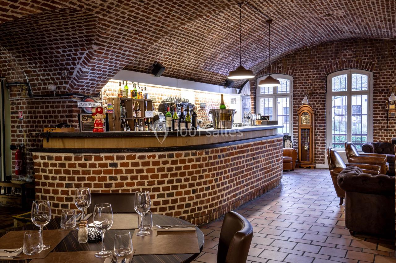 Bar en briques apparentes avec comptoir en bois, tables dressées au premier plan et fauteuils en cuir en arrière-plan.