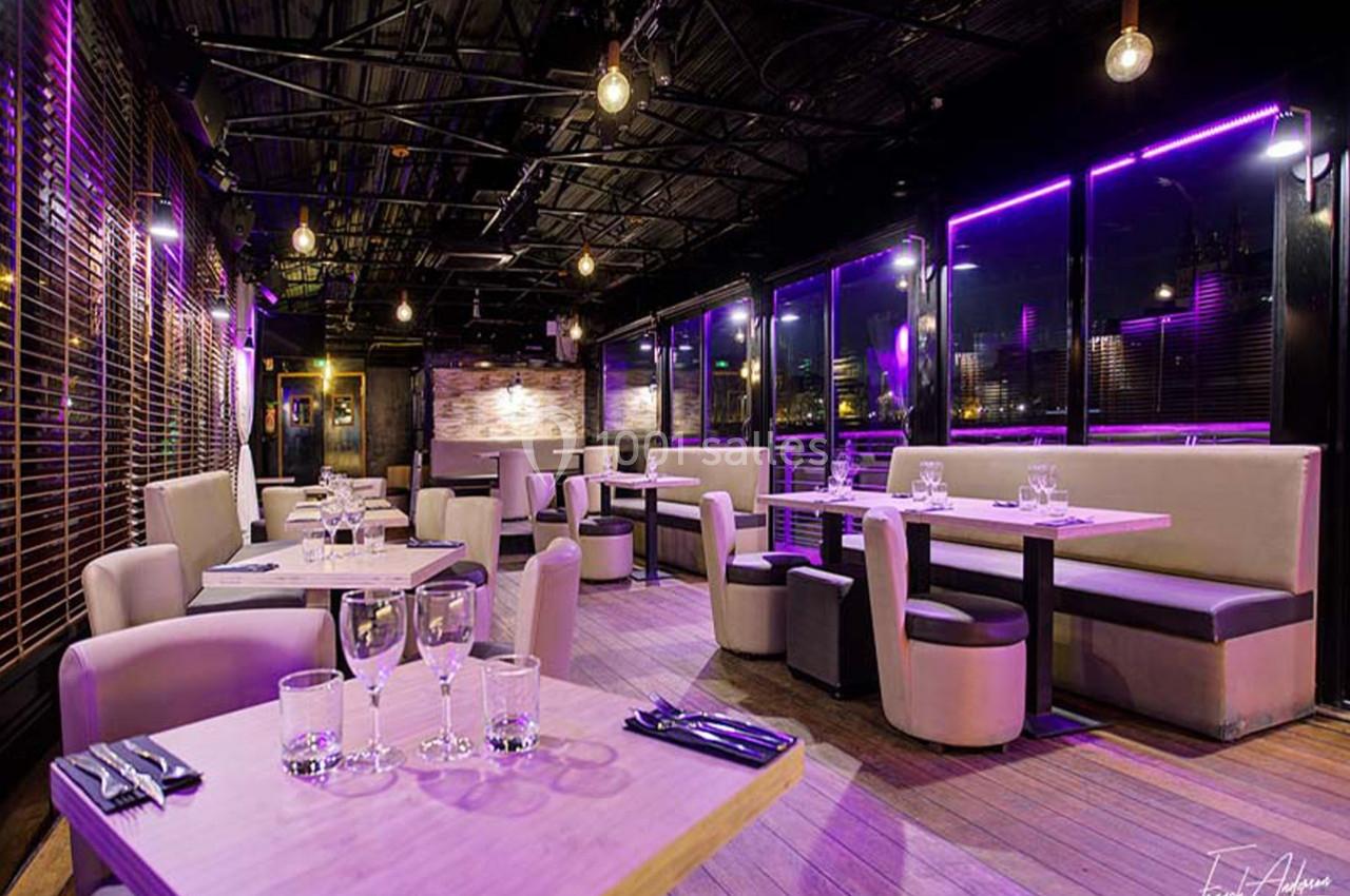 Salle de restaurant moderne avec éclairage violet, tables dressées et grandes baies vitrées donnant sur l'extérieur.