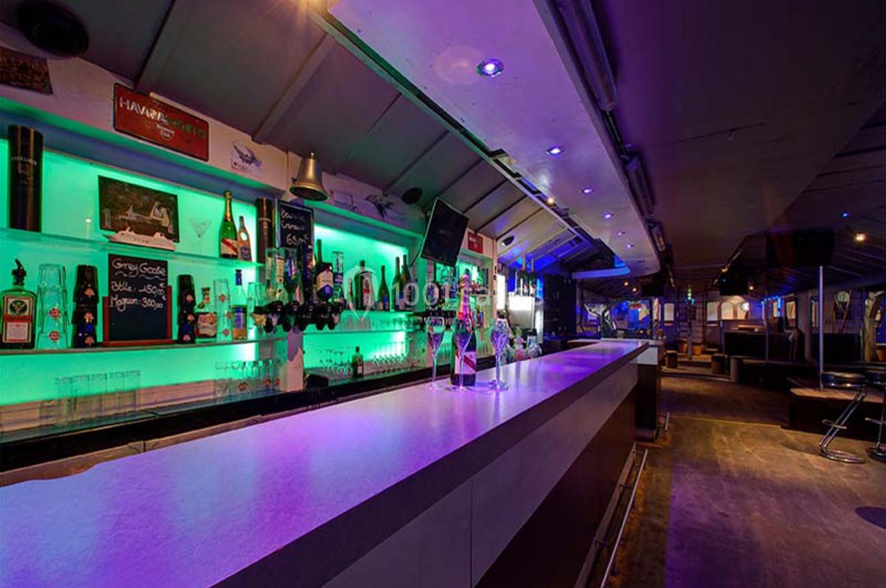 Bar moderne avec éclairage néon vert, comptoir long et rangées de bouteilles alignées sur des étagères.