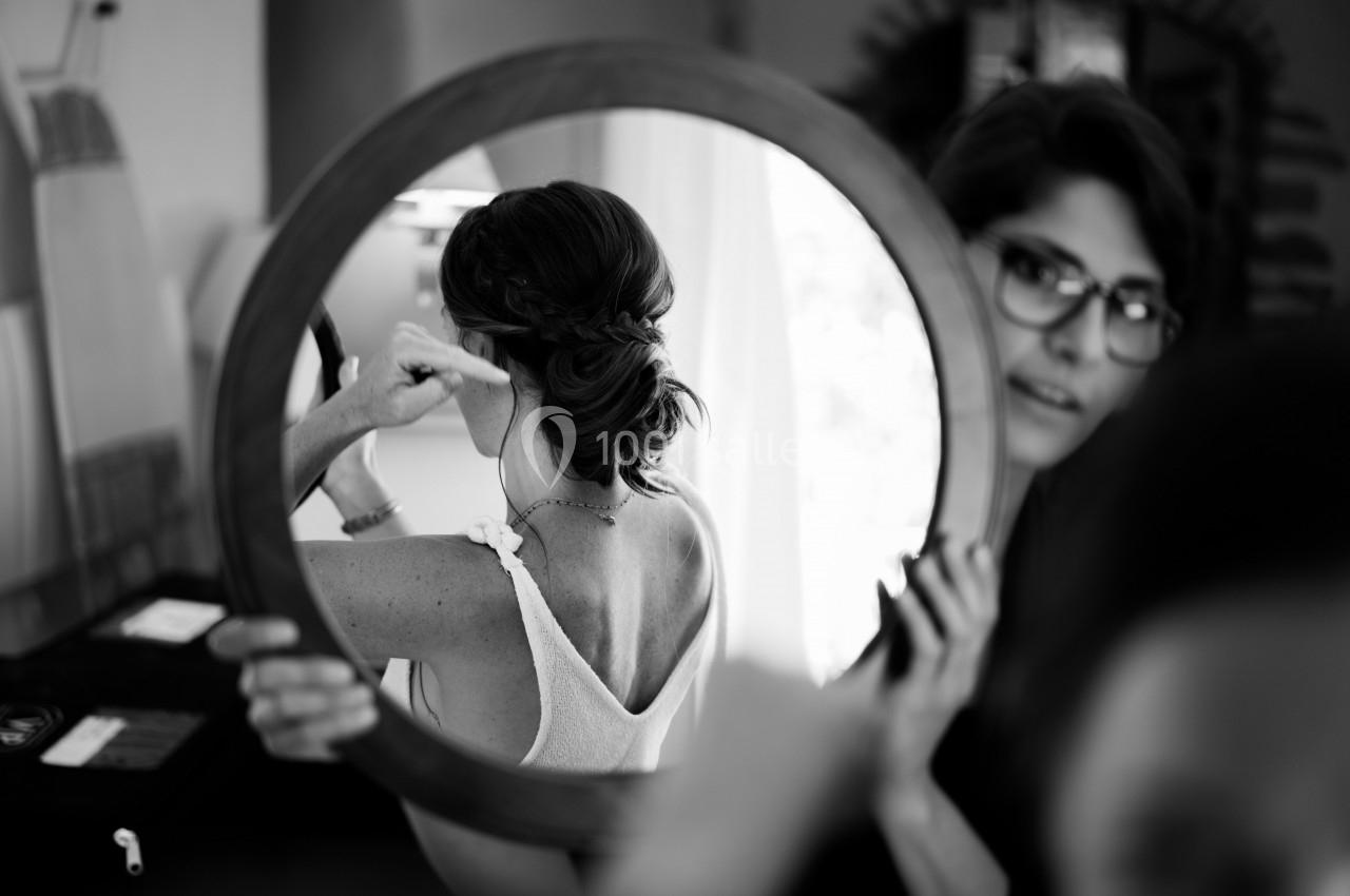 Une femme ajuste sa coiffure devant un miroir rond, reflétant son dos et son chignon tressé.