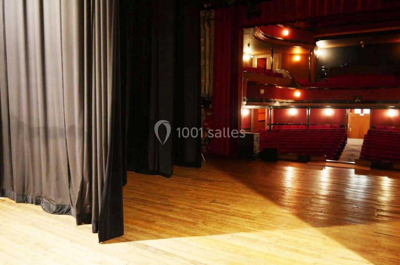 Vue depuis les coulisses d'une scène de théâtre avec rideaux noirs, plancher en bois et salle vide éclairée.