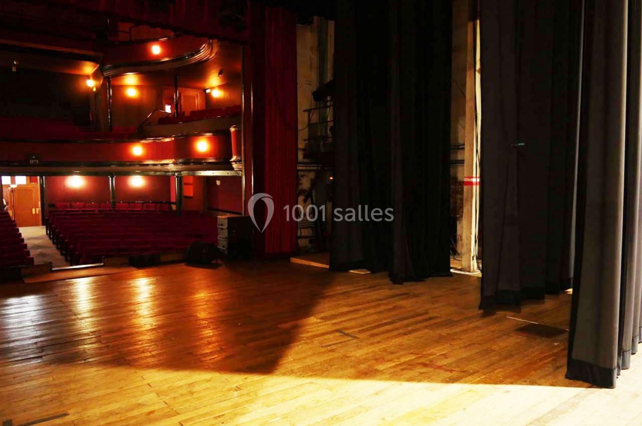 Vue depuis la scène d'un théâtre vide, montrant les sièges rouges, les rideaux noirs et l'éclairage tamisé.