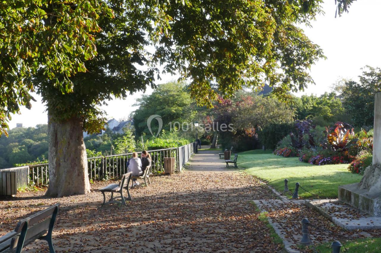Allée bordée d'arbres et de bancs dans un parc, avec des personnes assises et des massifs de fleurs en arrière-plan.