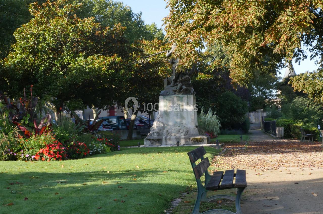 Statue entourée de verdure dans un parc, avec un banc en premier plan et des arbres ombrageant l'allée.