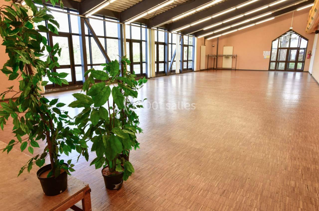 Grande salle lumineuse avec parquet, baies vitrées, poutres apparentes et deux plantes en pot au premier plan.