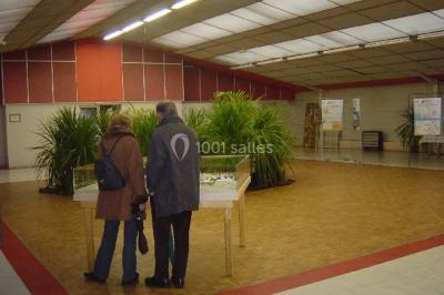 Deux personnes observent une maquette sous vitrine dans une salle avec des plantes et des panneaux d'exposition.