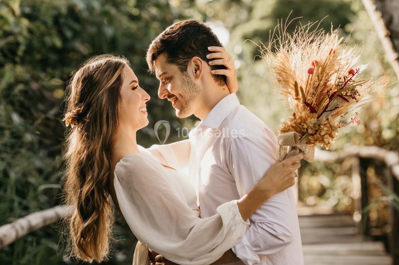 Un couple souriant s'enlace sur un pont en bois, la femme tenant un bouquet de fleurs séchées.