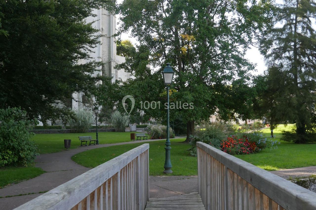 Passerelle en bois menant à un parc verdoyant avec bancs, lampadaire et arbres en arrière-plan.