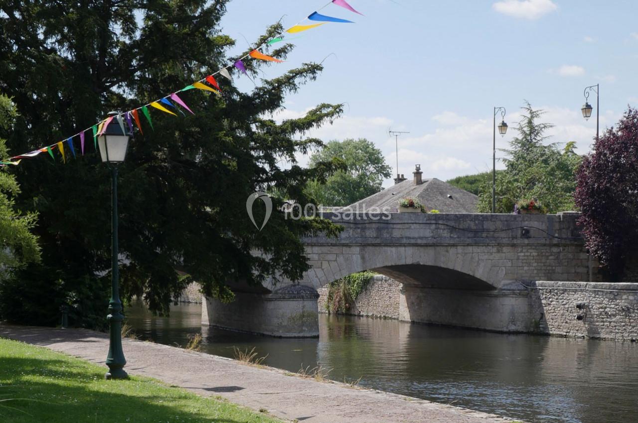 Pont en pierre traversant une rivière, entouré d'arbres, avec des fanions colorés suspendus et un lampadaire au premier plan.