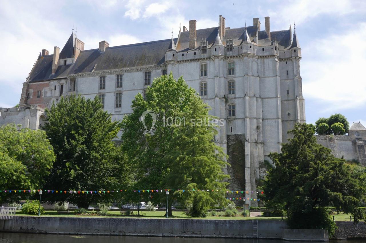 Façade d'un château médiéval en pierre, entouré d'arbres et bordé d'une rivière avec des guirlandes colorées.