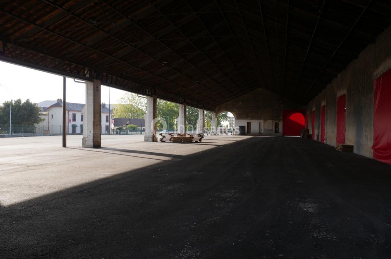 Hangar ouvert avec une structure métallique, sol asphalté, lumière naturelle et bâtiments visibles à l'extérieur.
