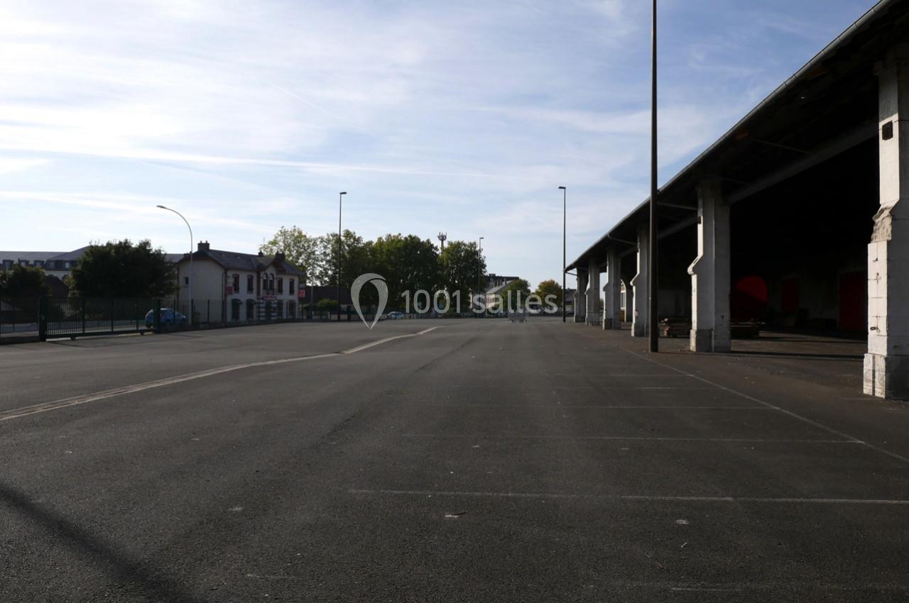 Grand parking vide avec un bâtiment couvert sur la droite et quelques arbres en arrière-plan sous un ciel dégagé.