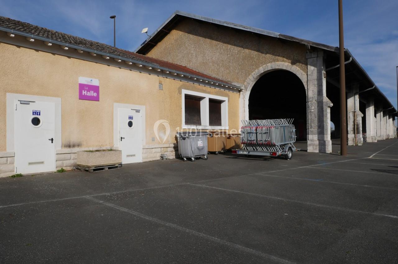 Bâtiment avec une grande arche, portes blanches, conteneurs et chariot de barrières sur un parking asphalté.