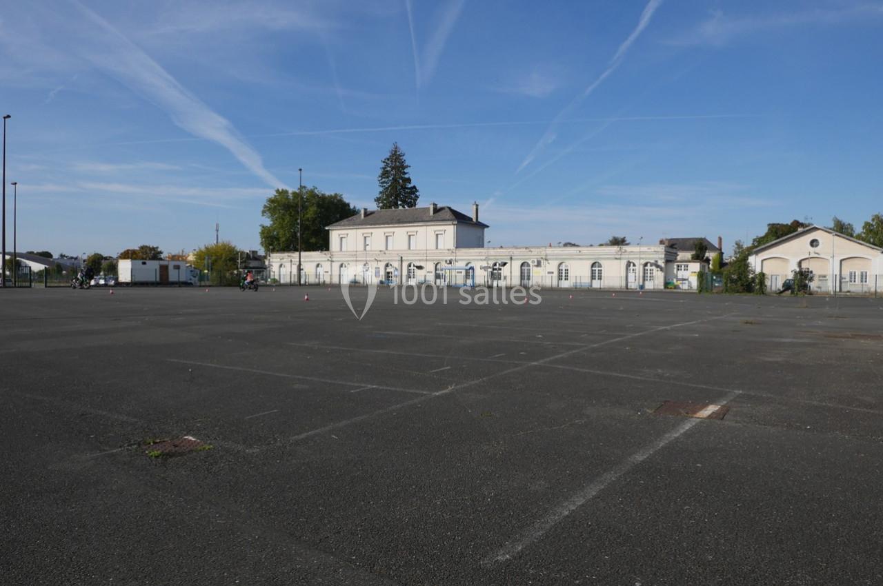 Grande place asphaltée vide devant un bâtiment blanc de style ancien, sous un ciel bleu traversé de traînées d'avions.