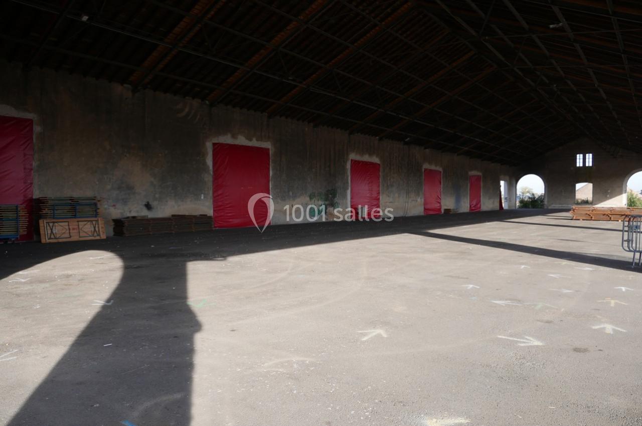 Intérieur d'un grand hangar vide avec des murs en béton, des portes rouges et un toit en charpente métallique.