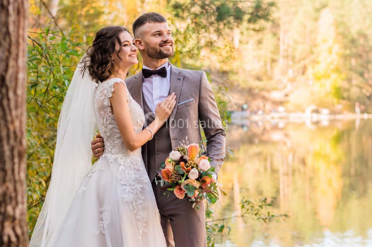 Un couple en tenue de mariage pose près d'un lac entouré de végétation automnale.