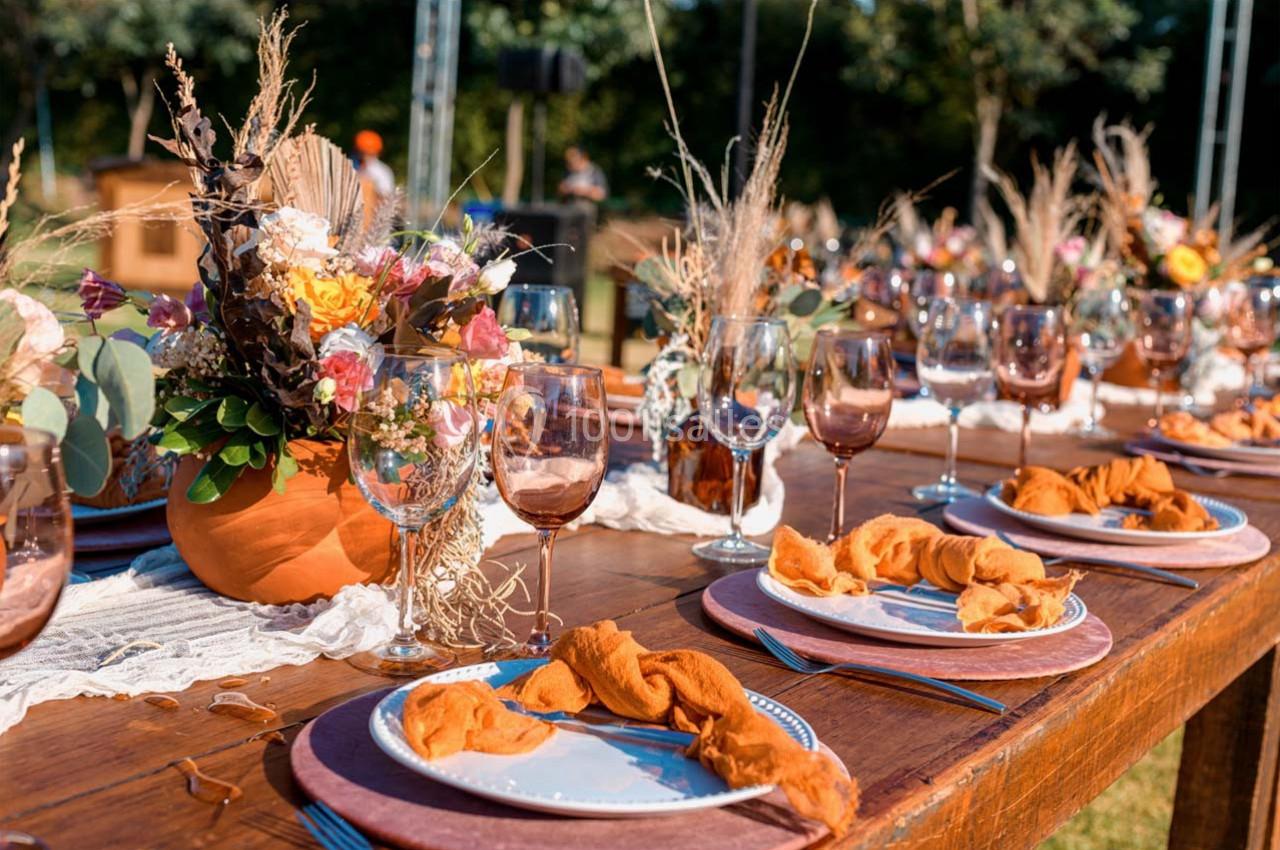 Table en bois décorée pour un repas en extérieur, avec fleurs séchées, verres colorés et serviettes orange.