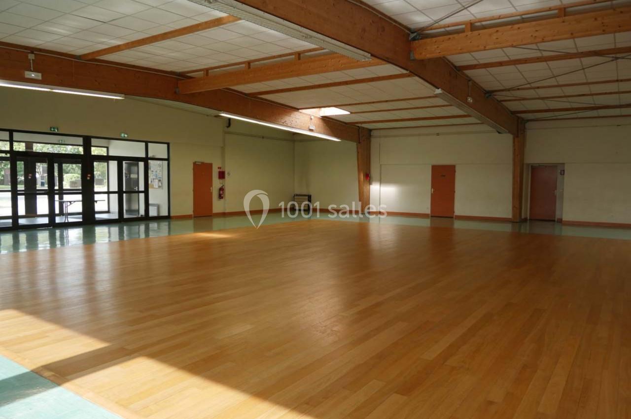 Salle vide avec parquet en bois clair, murs clairs, grandes baies vitrées et poutres apparentes au plafond.
