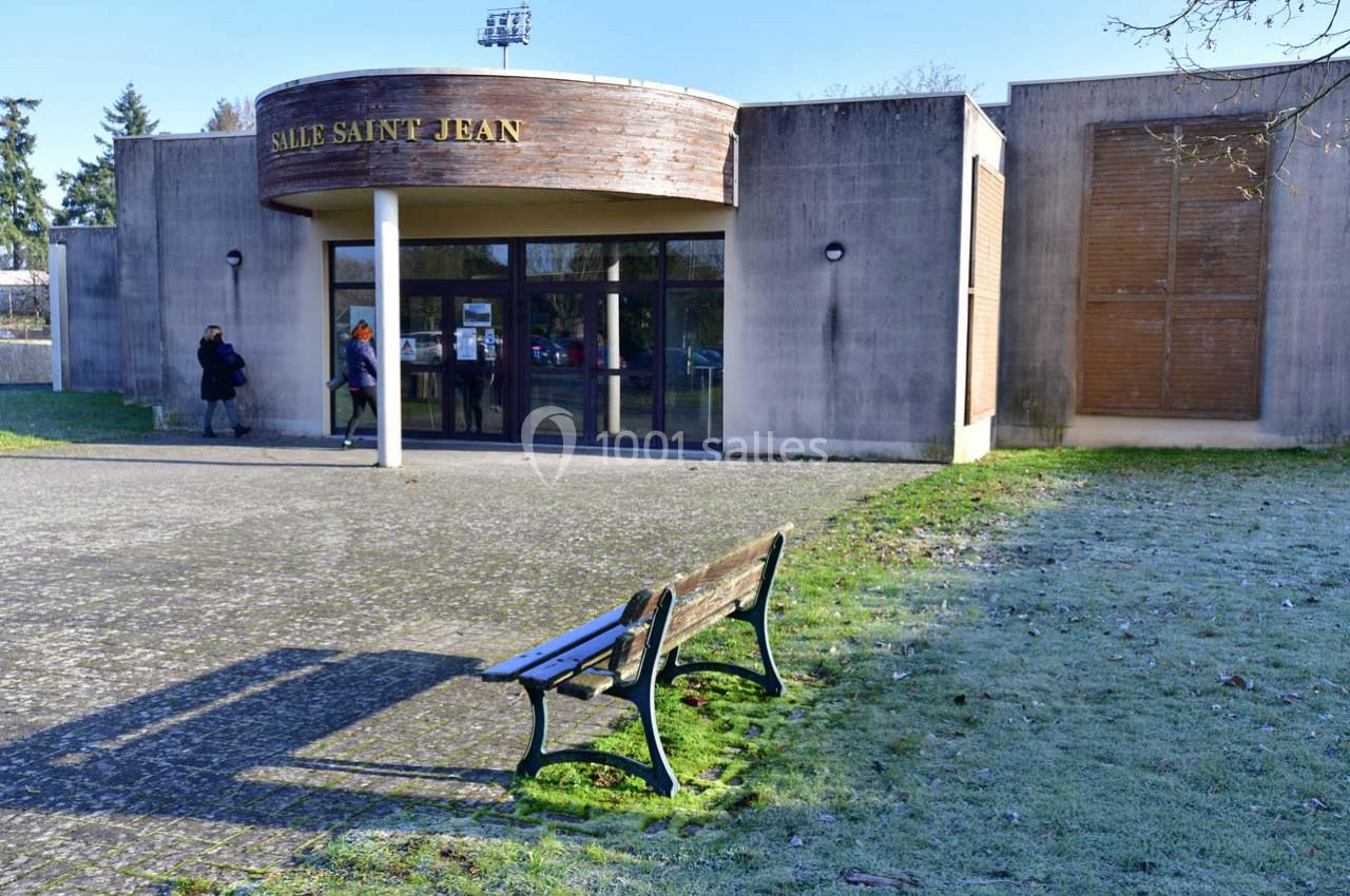 Façade d'un bâtiment nommé ’Salle Saint Jean’ avec une entrée vitrée, un banc et un espace extérieur herbeux.