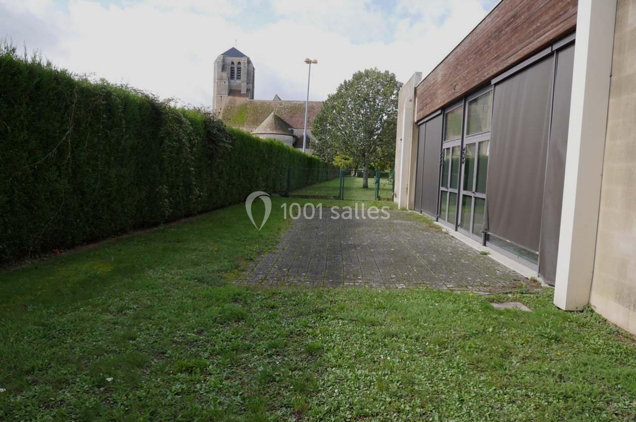 Allée pavée bordée de pelouse et de haies, menant à un bâtiment moderne avec une église en arrière-plan.