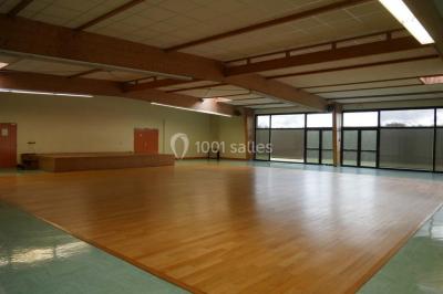 Salle vide avec parquet en bois, murs clairs, plafond avec poutres apparentes et grandes baies vitrées.