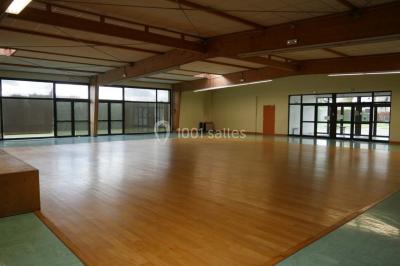 Salle vide avec parquet en bois, murs clairs, plafond avec poutres apparentes et grandes baies vitrées.