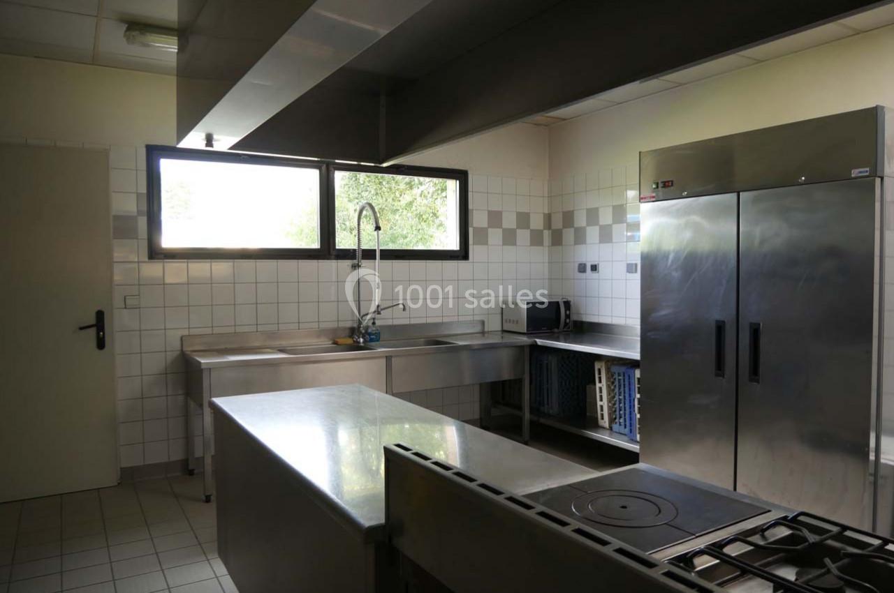 Cuisine professionnelle équipée avec plan de travail en inox, réfrigérateur, évier, plaques de cuisson et fenêtres.