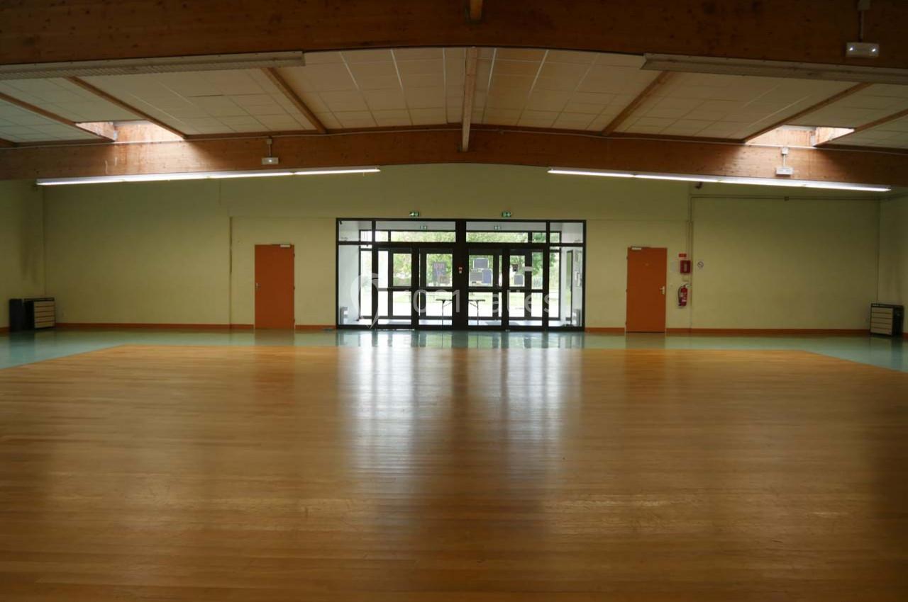 Salle vide avec parquet en bois, murs clairs, grandes fenêtres vitrées et plafond avec poutres apparentes.