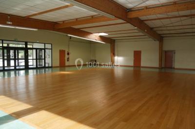 Salle vide avec parquet en bois, murs clairs, plafond avec poutres apparentes et grandes baies vitrées.