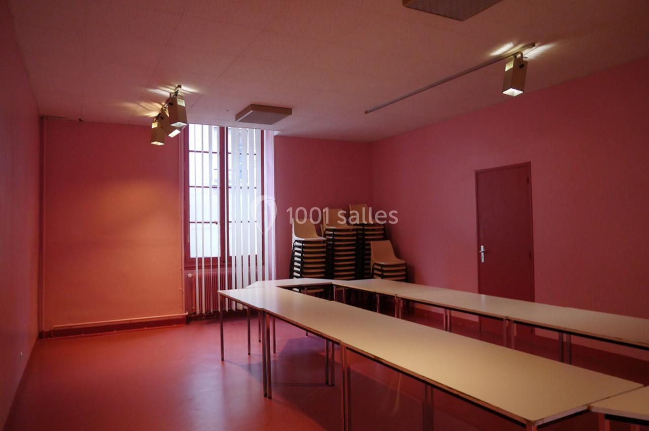 Salle vide avec murs roses, tables blanches alignées en U et chaises empilées près d'une fenêtre à barreaux.