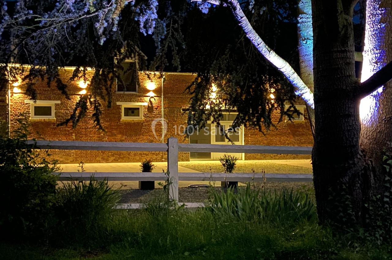 Façade en briques éclairée la nuit, entourée d'arbres et d'une clôture blanche au premier plan.