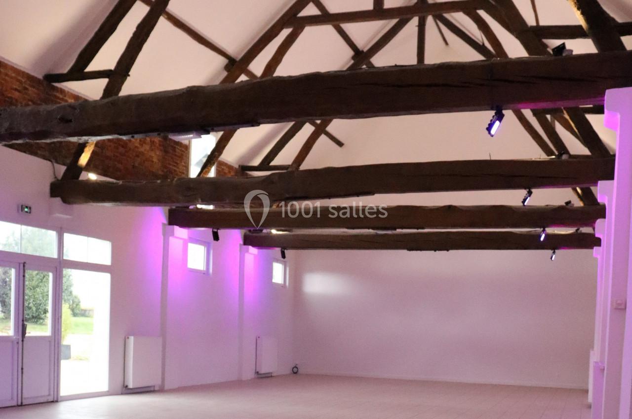 Salle spacieuse avec poutres apparentes, murs blancs, éclairage violet et carrelage au sol.