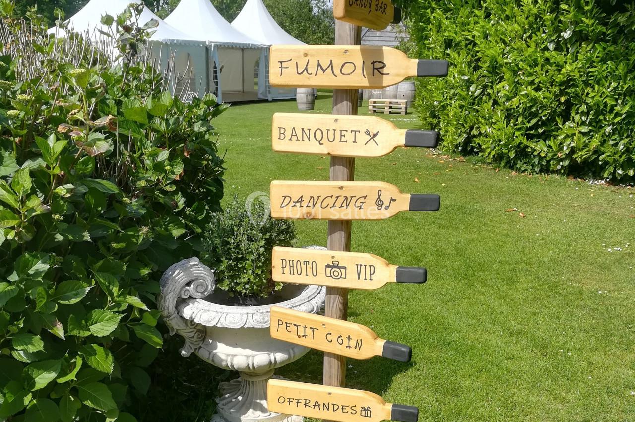 Panneau en bois avec flèches indiquant différentes zones d'un événement en plein air, entouré de verdure.