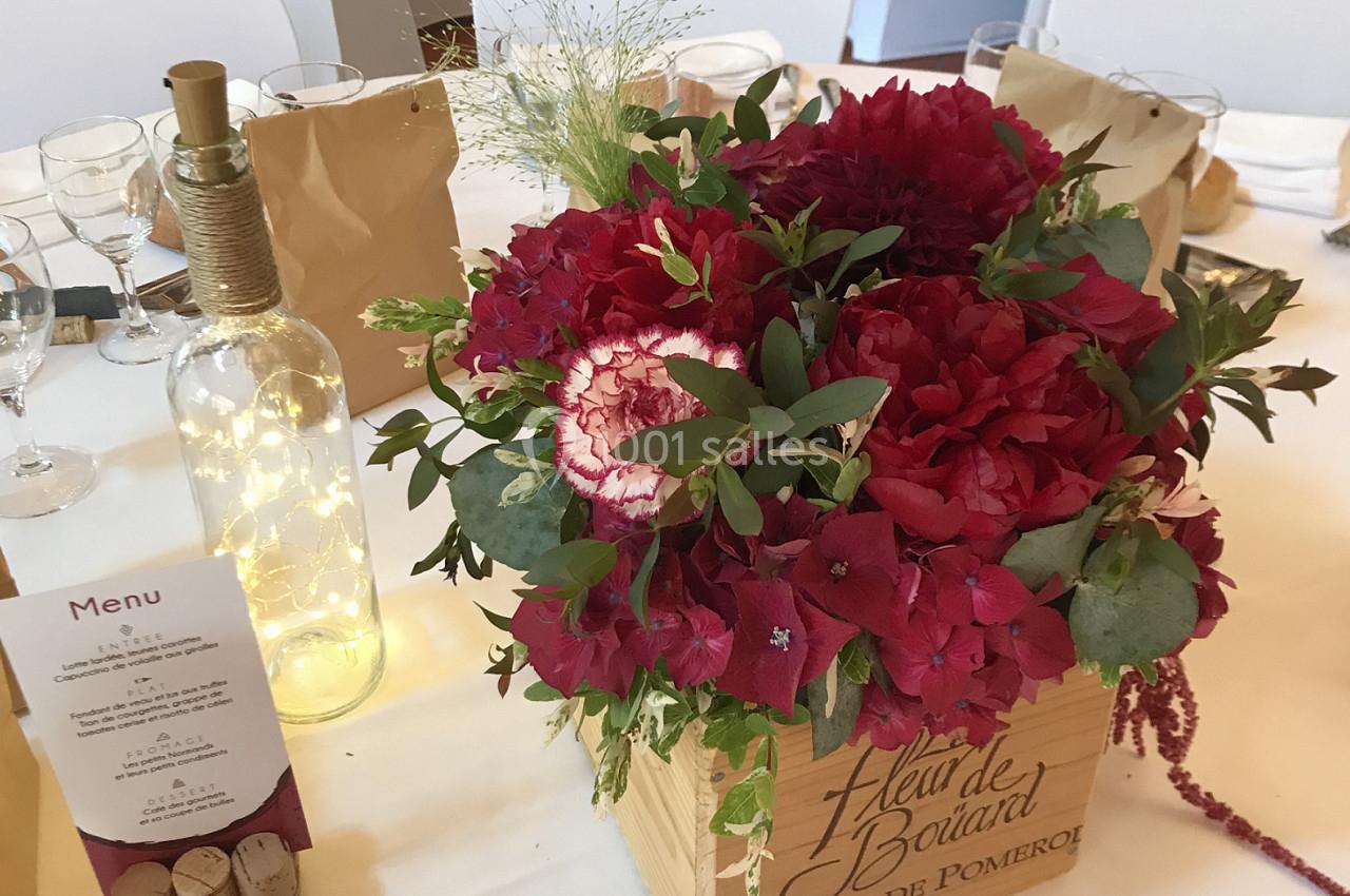 Centre de table avec fleurs rouges dans une caisse en bois, bouteille décorative lumineuse et menu sur une table dressée.
