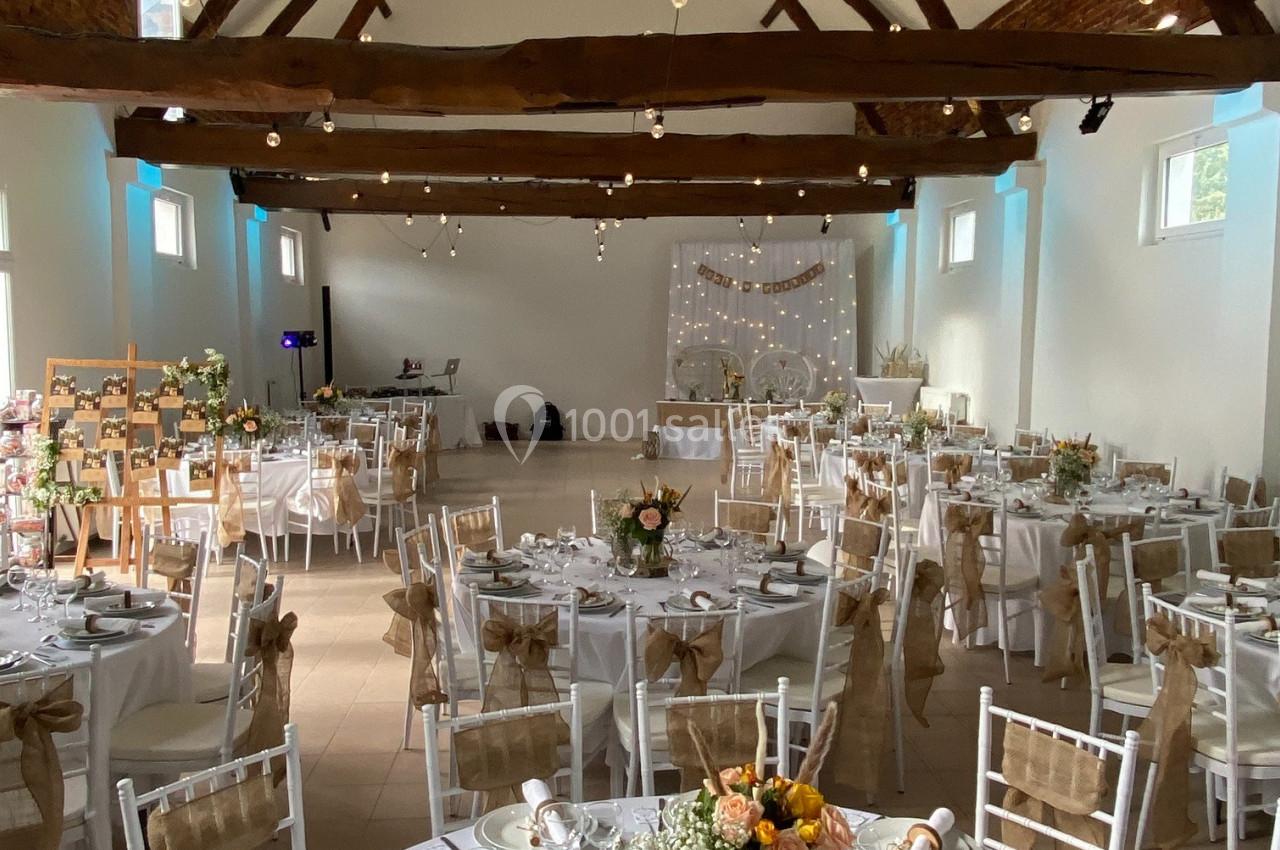 Salle de réception décorée pour un mariage, avec tables dressées, chaises blanches et poutres apparentes au plafond.