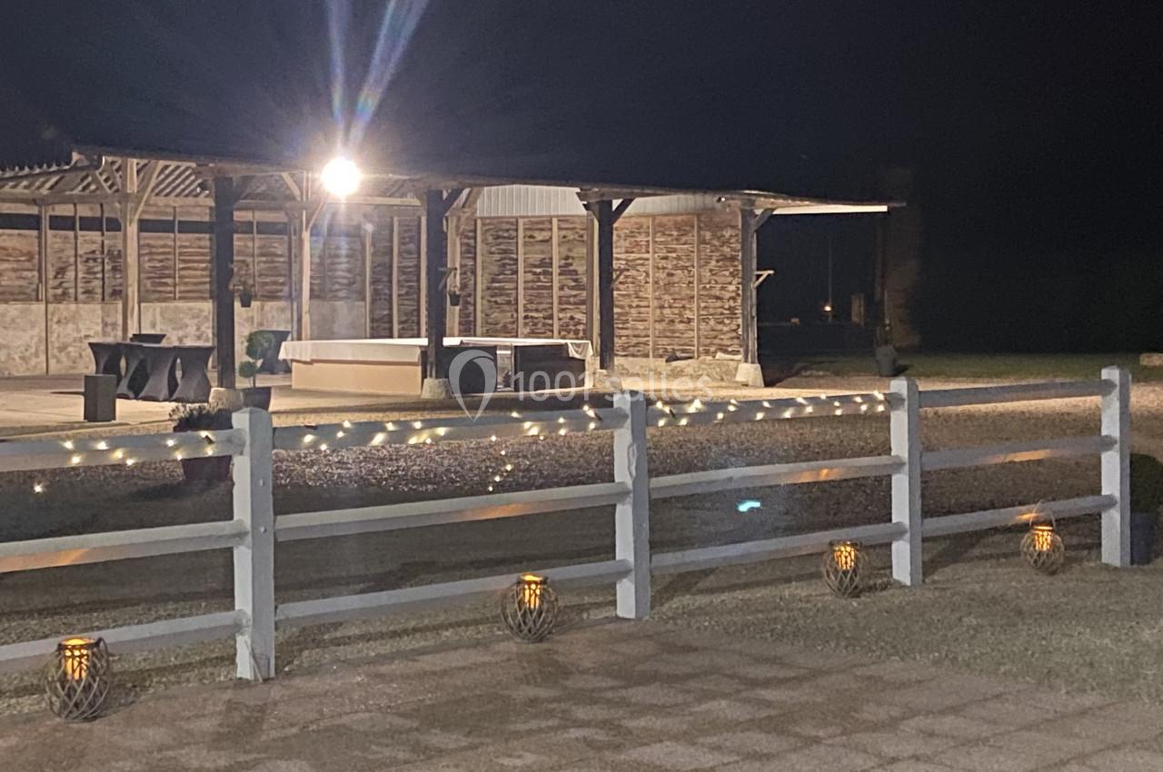 Espace extérieur éclairé de nuit avec une pergola, des guirlandes lumineuses et des lanternes au sol.