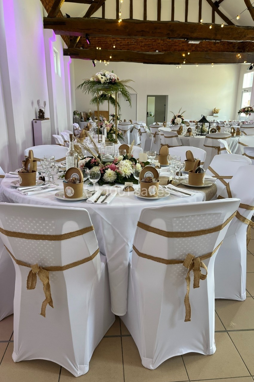 Salle de réception décorée pour un mariage, avec tables rondes dressées, nappes blanches et ornements floraux.