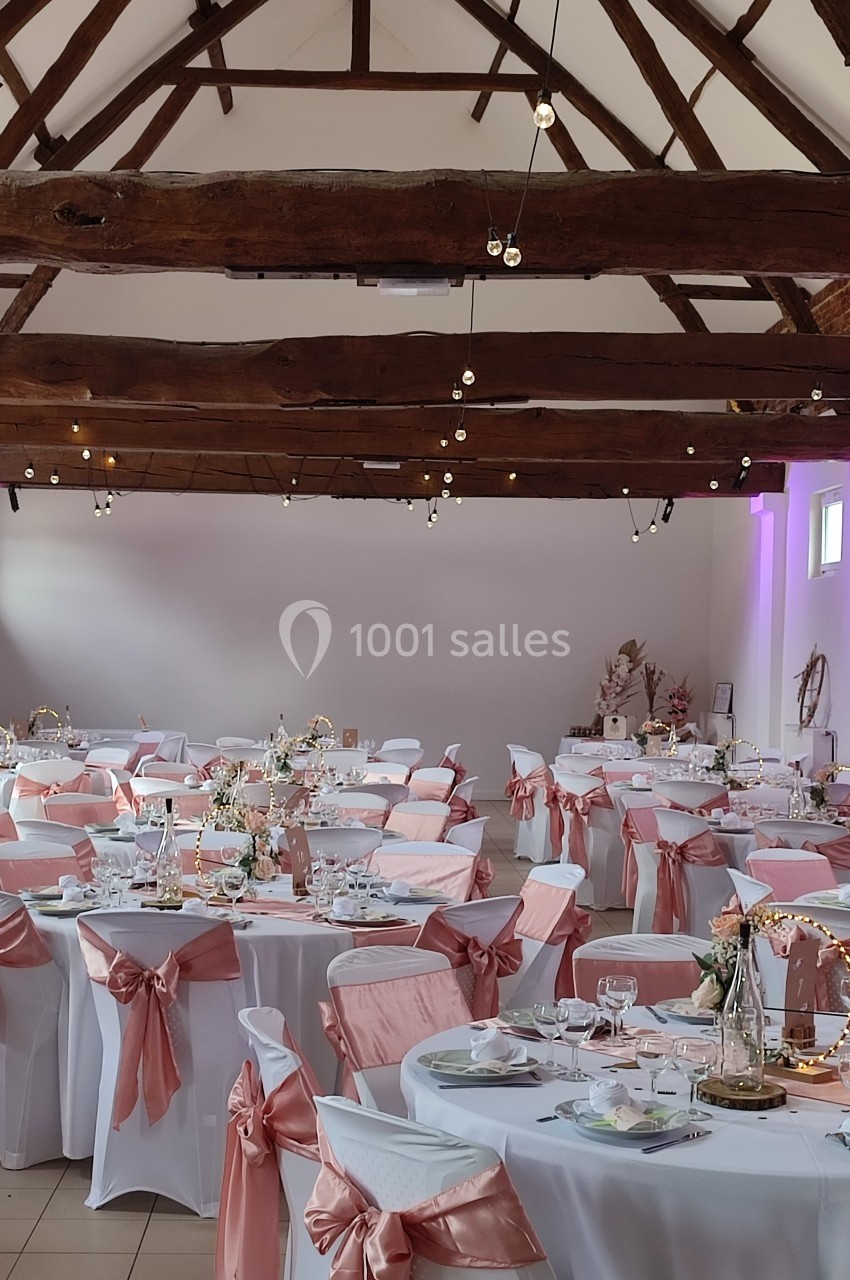 Salle de réception décorée pour un mariage, avec des tables dressées et des chaises ornées de nœuds roses.