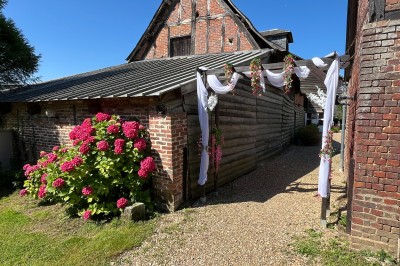 Allée en gravier bordée d'un bâtiment en bois et briques, décorée de fleurs et de voilages blancs.
