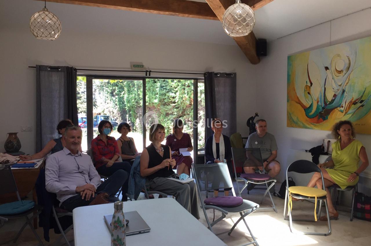 Un groupe de personnes assises dans une salle lumineuse, attentives, avec des tables et un tableau coloré en arrière-plan.