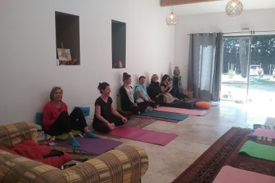 Des personnes assises sur des tapis et des chaises dans une salle lumineuse avec un tableau et des rideaux.