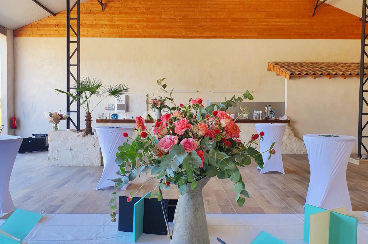 Salle lumineuse avec des tables hautes drapées de blanc et un grand bouquet de fleurs roses au premier plan.