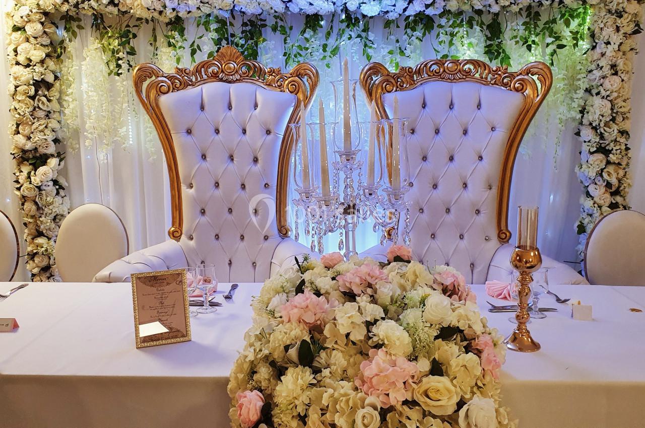 Deux fauteuils ornés dorés devant un mur de fleurs blanches, avec une table décorée de bougies et de fleurs.