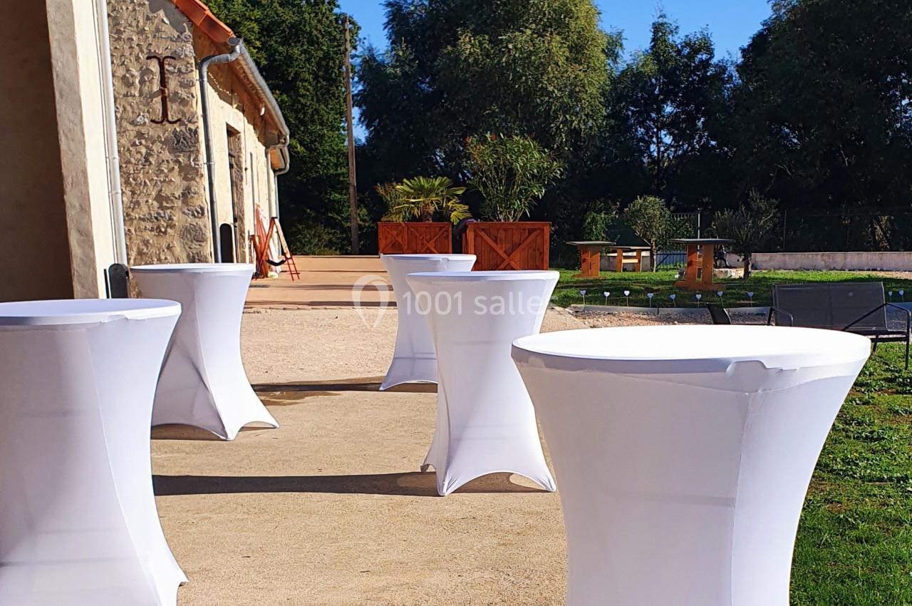 Tables hautes recouvertes de housses blanches disposées sur une terrasse ensoleillée près d'un bâtiment en pierre.
