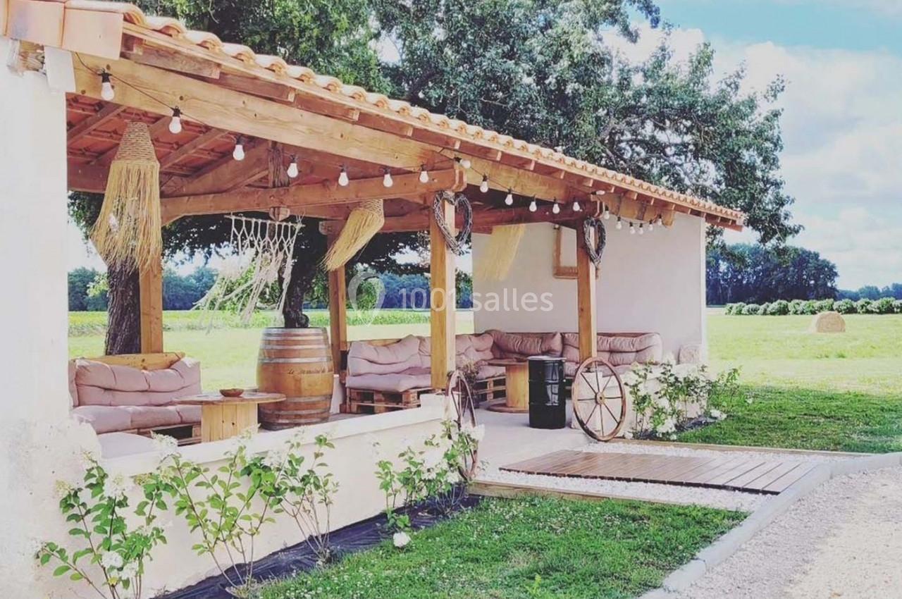 Terrasse couverte en bois avec canapés, décor champêtre, tonneau en table et vue sur un champ verdoyant.