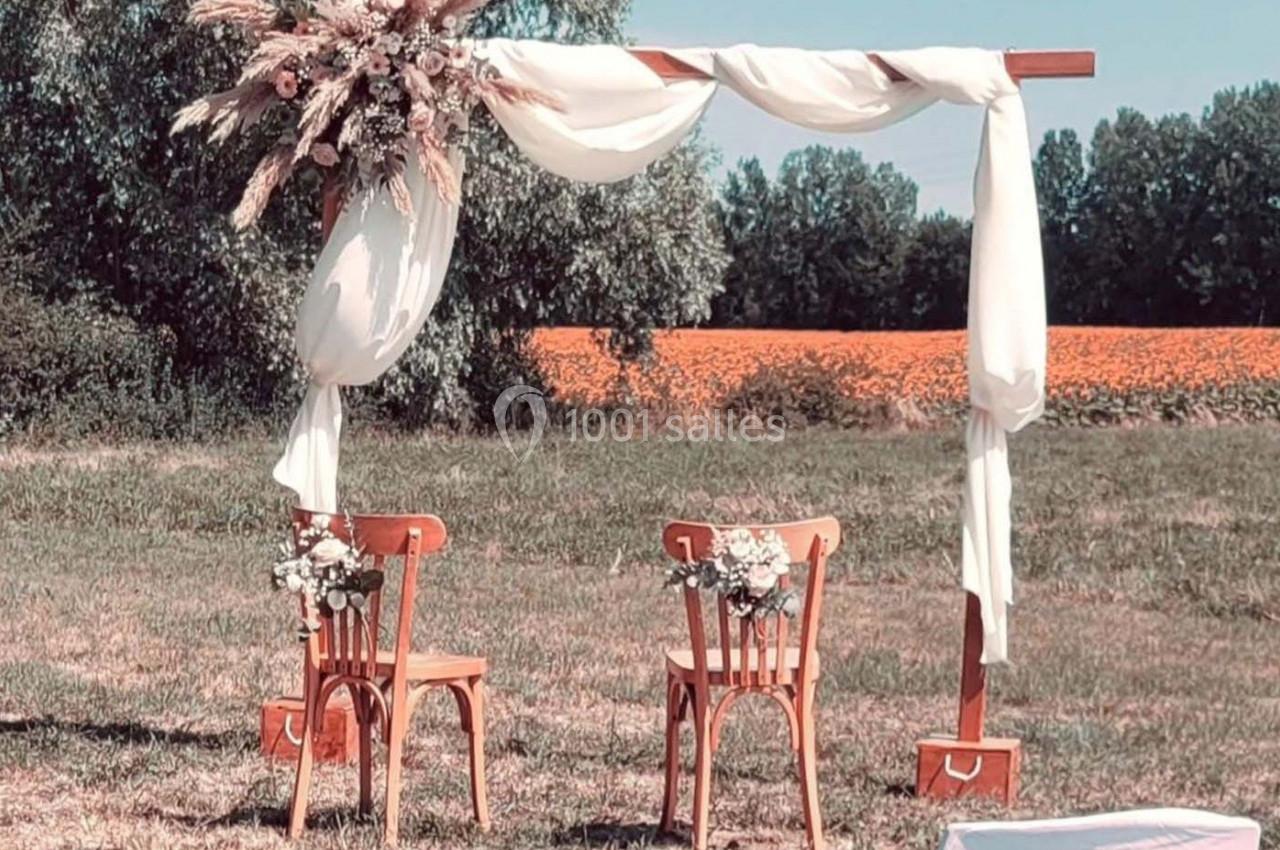 Arche de mariage en bois décorée de fleurs séchées et de voilages blancs, avec deux chaises sur une pelouse en plein air.
