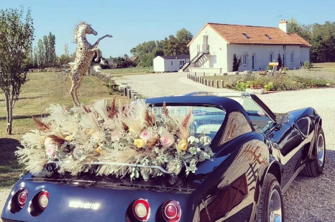 Voiture décapotable noire décorée de fleurs séchées, stationnée devant une maison de campagne avec jardin.